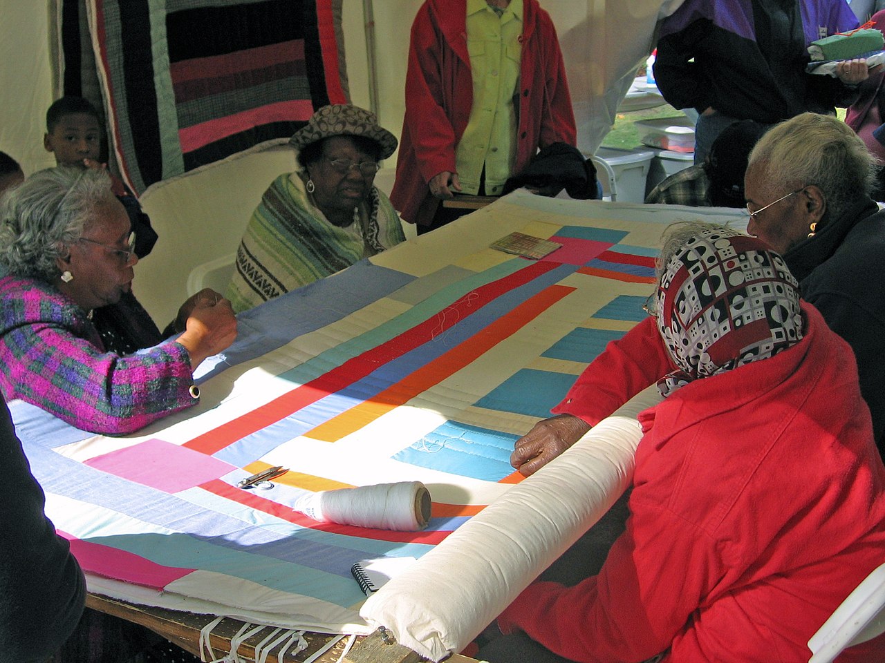 Women sitting together and sewing a big, colorful quilt by hand.