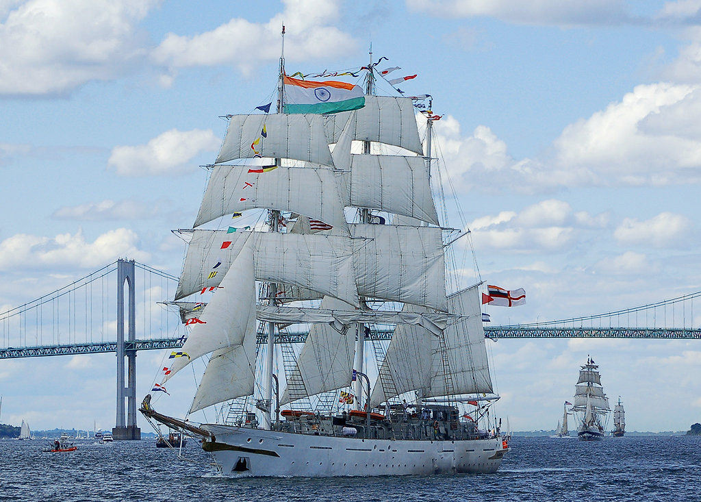 Tall sailing ships glide across the bay under a big bridge.