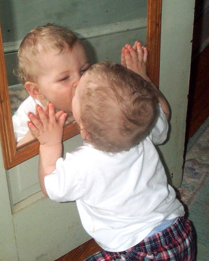 A smiling baby gives a kiss to its own mirror reflection.