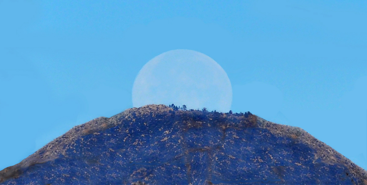 A soft full moon setting behind a tall mountain at sunrise.